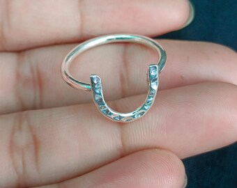 May include: A silver-colored ring featuring a horseshoe-shaped focal point. The horseshoe element has a textured, hammered surface finish, while the band of the ring is smooth and circular. The metal has a polished, reflective appearance throughout.