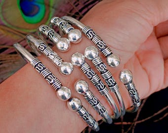 May include: A collection of silver-toned metal bangles. Each bangle features a decorative design with a central beaded element and etched patterns. The bracelets are displayed on a wrist, showcasing their intricate details.