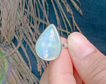 May include: A silver ring featuring a teardrop-shaped, iridescent moonstone cabochon. The gemstone displays a milky white appearance with subtle blue light reflections. The stone is set in a smooth, polished metal bezel on a simple band.