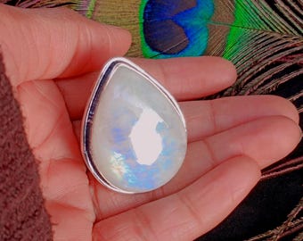 May include: A pear-shaped, iridescent moonstone pendant with a silver-colored bezel. The stone displays a milky white base with flashes of blue and rainbow hues. The pendant is held in an open hand, with a peacock feather in the background.