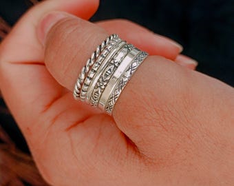 May include: A hand wearing a stack of silver rings. The rings feature various designs, including twisted rope, etched patterns, and geometric shapes. The rings are worn on a finger, showcasing the different textures and styles.