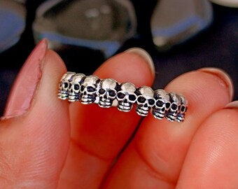 May include: A silver ring featuring a repeating pattern of small skull designs. The ring is held between fingers, showcasing the detailed craftsmanship of the skull motifs. The skulls are evenly spaced around the band.