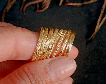 May include: A collection of gold-colored rings, each with a unique textured design. The rings are stacked together, showcasing various patterns, including floral and beaded details. The rings are held by a person's fingers.