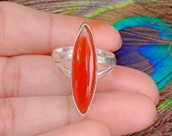 May include: A silver ring featuring a vibrant, elongated, oval-shaped, red gemstone. The ring's band has a split design, adding a unique detail to the jewelry. The gemstone is the focal point, showcasing a polished, smooth surface.