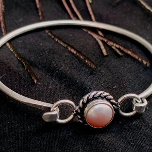 Peut inclure: Un bracelet jonc en argent avec un accent de perle centrale. La perle est sertie dans un cadre circulaire texturé, relié au bracelet par de petits maillons décoratifs. Le bracelet est sur fond sombre.