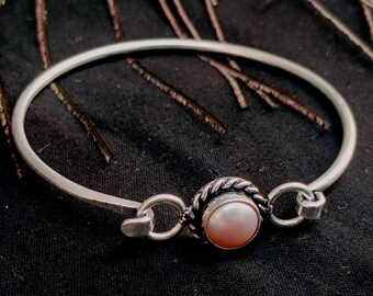 May include: A silver bangle bracelet with a central pearl accent. The pearl is set in a textured, circular frame, connected to the bracelet with small, decorative links. The bracelet is on a dark background.