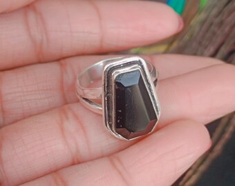 May include: A silver ring featuring a large, rectangular black gemstone. The gemstone is faceted, catching the light and creating a striking contrast against the silver band. The ring's design is simple yet elegant, suitable for everyday wear or special occasions.