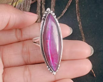 May include: A silver ring featuring a large, elongated, oval-shaped, polished gemstone in shades of pink and purple. The gemstone is set in a detailed silver bezel with a beaded border.