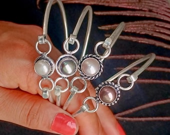 May include: Four silver-toned bangles, each featuring a central pearl or pearl-like stone. The pearls vary in color, from white to grey and pink. The bangles are connected with small silver rings.