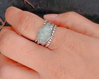 May include: A silver ring featuring a large, irregular-shaped, pale blue-green gemstone. The ring has a textured band with small, round embellishments. The gemstone is set in a silver bezel, creating a unique and naturalistic design.