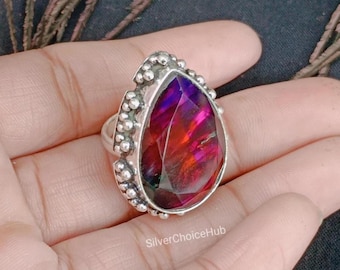 May include: A sterling silver ring featuring a large, teardrop-shaped gemstone with vibrant red, purple, and orange hues. The gemstone is set in a detailed bezel with small, beaded accents. The ring is displayed on a hand.