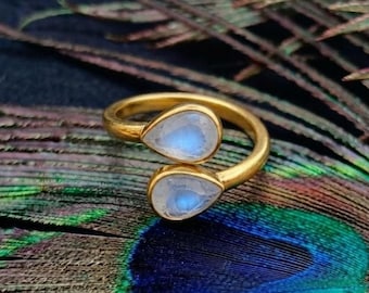 May include: A gold-toned open ring featuring two teardrop-shaped moonstones. Each stone displays a soft, iridescent blue glow set within a smooth metal bezel. The band is polished and curves to create a bypass design with the stones facing each other.