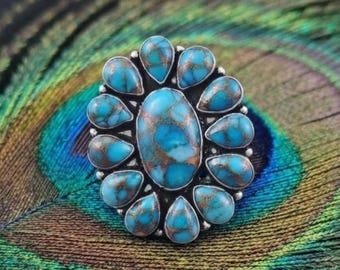 May include: A silver ring featuring a large oval turquoise stone in the center, surrounded by twelve smaller teardrop-shaped turquoise stones. Each stone displays a vibrant blue color with intricate brown matrix veining throughout the surface.