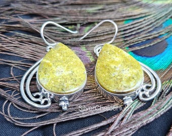 May include: A pair of silver earrings with teardrop-shaped, yellow-green gemstones. The earrings have a decorative silver design around the gemstone and a hook closure. The earrings are displayed on a background of peacock feathers.