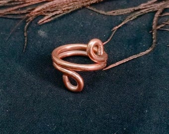 May include: A copper ring crafted from a single continuous wire, featuring a double-loop design where the ends of the wire are coiled into small, rounded spirals. The metal has a polished, warm reddish-brown metallic finish and a smooth texture.