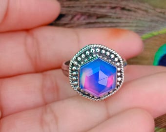 May include: A silver ring featuring a hexagonal, iridescent stone with pink and blue hues. The stone is set within a decorative, beaded silver border that frames the central gem, creating a textured, metallic edge around the faceted surface.