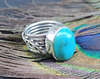 May include: A silver ring with a large, oval turquoise stone. The ring has a multi-band design with leaf-like detailing on the side. The turquoise stone is a bright, vibrant blue. The ring is displayed on a background of peacock feathers.