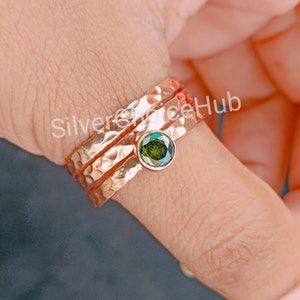 Pode incluir: Um conjunto de três anéis de ouro rosa martelado com uma gema verde no centro. Os anéis estão empilhados em um dedo.