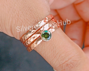 May include: A set of three hammered rose gold rings with a green gemstone in the center. The rings are stacked on a finger.