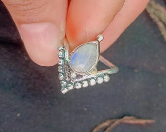 May include: A silver ring featuring a pear-shaped moonstone cabochon set in a V-shaped band. The metal band is decorated with a series of small, raised silver spheres along the edges, framing the central iridescent stone with a polished finish.