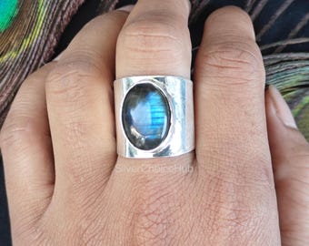 May include: A wide, silver-colored ring with an oval, iridescent blue gemstone. The ring has a thick band and a polished finish. The gemstone displays shades of blue and green. The ring is a piece of jewelry.