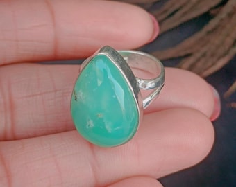 May include: A silver ring featuring a large, teardrop-shaped, light-green gemstone. The gemstone is set in a silver bezel and has a smooth, polished surface. The ring's band is split on either side of the gemstone.