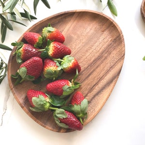 May include: A wooden serving platter with a natural wood grain finish, filled with fresh red strawberries. The strawberries are arranged in a cluster, with their green leaves visible.