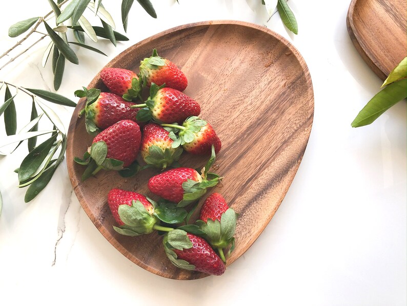 May include: A wooden serving platter with a natural wood grain finish, filled with fresh red strawberries. The strawberries are arranged in a cluster, with their green leaves visible.