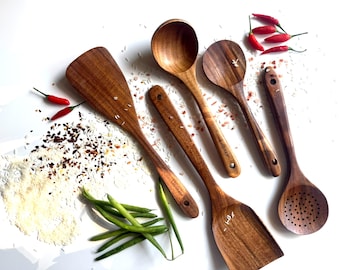 Puede incluir: Un conjunto de utensilios de cocina de madera, incluyendo una espátula y un cucharón, dispuestos sobre una superficie blanca. Chiles rojos, judías verdes y arroz se encuentran dispersos alrededor de los utensilios.