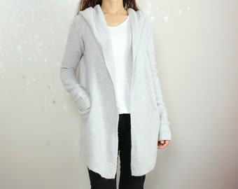 May include: A long, light gray hooded cardigan with a relaxed fit. The cardigan is open in the front and has pockets on the sides. The model is wearing black jeans and tall black boots.