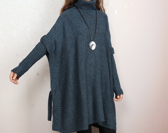 May include: A dark blue turtleneck sweater with long sleeves and a loose fit. The sweater has a tie detail at the sides and a silver pendant necklace.