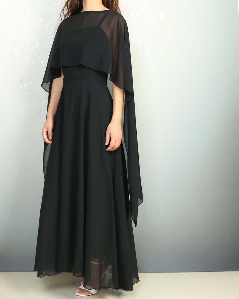 K&ouml;nnte beinhalten: Ein schwarzes Maxikleid mit einem transparenten Chiffon-Cape-&Uuml;berwurf. Das Kleid hat ein figurbetontes Oberteil und einen flie&szlig;enden Rock.