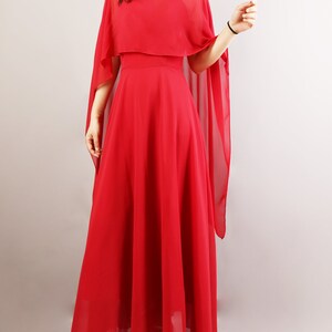 K&ouml;nnte beinhalten: Ein langes, rotes, flie&szlig;endes Maxikleid mit einem durchsichtigen Umhang dar&uuml;ber. Das Kleid hat ein tailliertes Oberteil und einen flie&szlig;enden Rock.