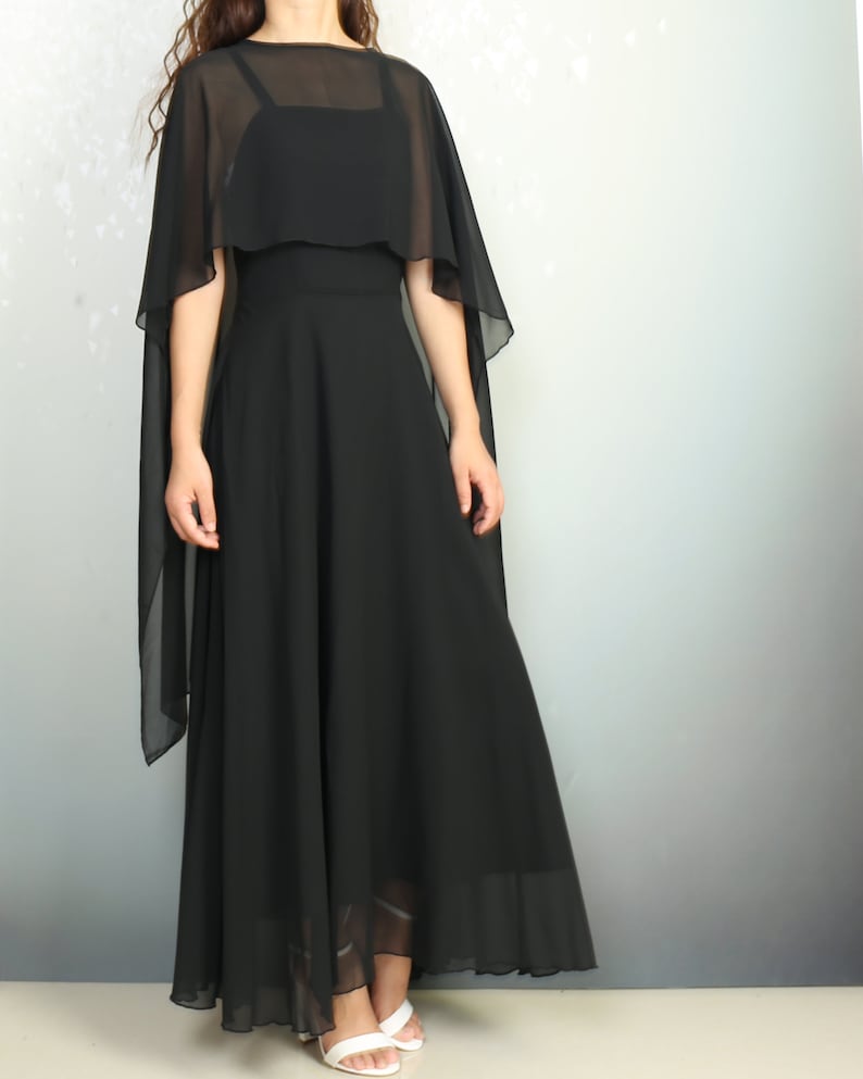K&ouml;nnte beinhalten: Ein schwarzes Maxikleid mit einem transparenten Chiffon-Cape-&Uuml;berwurf. Das Kleid hat ein figurbetontes Oberteil und einen flie&szlig;enden Rock.