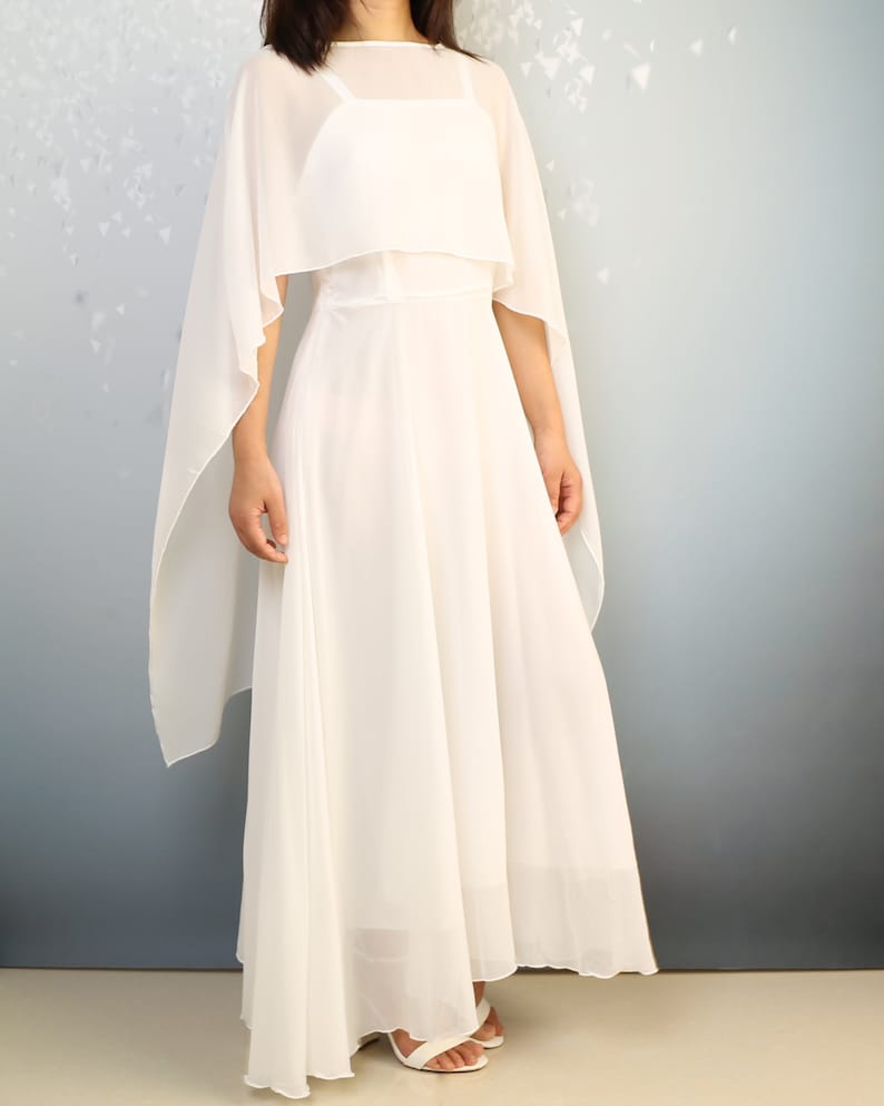 K&ouml;nnte beinhalten: Ein wei&szlig;es Maxikleid mit einem durchsichtigen Umhang dar&uuml;ber. Das Kleid hat ein tailliertes Oberteil und einen flie&szlig;enden Rock.