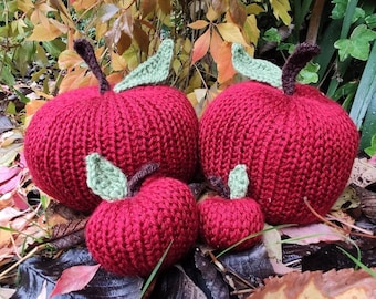 Op de afbeelding: Vier gehaakte appels in verschillende maten, in rode kleur met groene bladeren. De appels zijn gerangschikt op een bed van gevallen herfstbladeren.