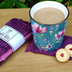 Puede incluir: Un posavasos de punto morado y rosa con una etiqueta blanca que dice "100% hecho a mano MUG RUG". El posavasos está sobre una superficie de madera con una taza con estampado floral y dos galletas con corazones rojos en la parte superior.