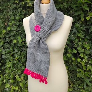 Puede incluir: Bufanda de punto gris con un botón rosa y borlas rosas en los extremos. La bufanda se muestra en un maniquí blanco.