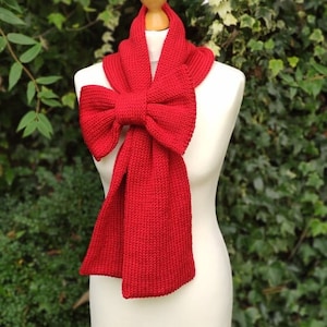 Pode incluir: Uma echarpe de malha vermelha com um grande laço amarrado na frente. A echarpe é exibida em um manequim branco contra um fundo verde de folhas.