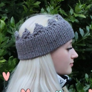 May include: A gray knitted crown headband with a pointed top. The headband is worn by a person with long blonde hair. The background is a green bush with colorful hearts.