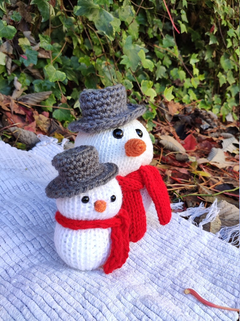 May include: Two crocheted snowmen, one larger than the other, with gray hats and red scarves. The snowmen are standing on a white surface.