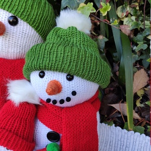 May include: Three crocheted snowmen with red and green hats and scarves. The snowmen are standing in front of a green leafy background.