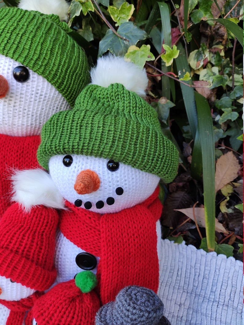 May include: Three crocheted snowmen with red and green hats and scarves. The snowmen are standing in front of a green leafy background.