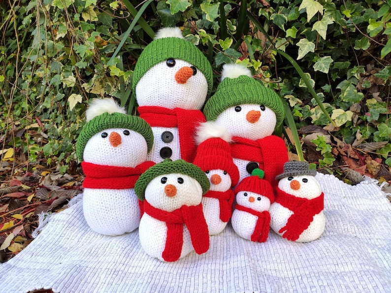 May include: A group of seven knitted snowman ornaments with green hats and red scarves. The snowmen are arranged in a row on a white knitted surface.