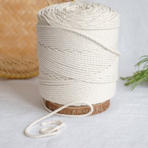 May include: A spool of natural white cotton rope, wound around a wooden base. The rope is thick and textured, and the spool is sitting on a white surface.