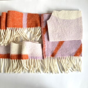 May include: A woven scarf with a cream-coloured fringe. The scarf features orange and lavender stripes and blocks. The scarf is made of a soft, textured material. The scarf is laid out on a white surface.