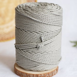 Könnte beinhalten: Eine große Spule grauer Makramee-Kordel, fest gewickelt und auf einer kleinen Holzscheibe präsentiert. Die Kordel hat eine neutrale Farbe und eignet sich für verschiedene Bastelprojekte. Der Hintergrund umfasst einen geflochtenen Korb und Grünzeug.