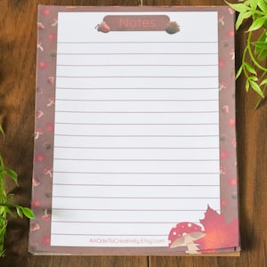 May include: A lined notepad with a brown border featuring mushrooms and leaves. The top of the notepad reads "Notes" and the bottom right corner has a mushroom and leaf design. The notepad is on a wooden surface.