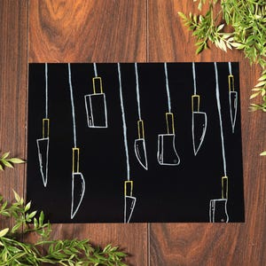 May include: A black art print with white line drawings of knives suspended from white lines. The knife handles are outlined in yellow. The print is set against a dark background and is framed by green foliage.