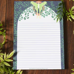 May include: Lined notepad with a decorative border featuring a green and white design. The top of the notepad has a green and yellow moth illustration. The notepad is on a wooden surface, with green leafy plants in the background.